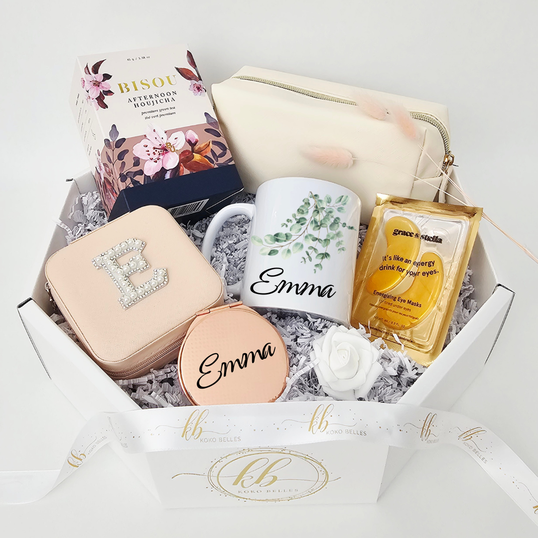A luxury personalized birthday care package for her by Koko Belles, highlighting a custom ceramic mug decorated with an elegant green eucalyptus vine design and the name Emma. The high-end octagonal gift box is styled with a white rose, cream pouch, pearl initial jewelry case, and a gold-wrapped eye mask.
