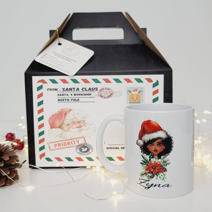 A white ceramic mug with a festive Christmas illustration of a woman in a Santa hat and the name 'Egna' printed on it, displayed in front of its black gift box with a holiday-themed label. The scene is styled with pinecones and warm fairy lights on a clean white background.