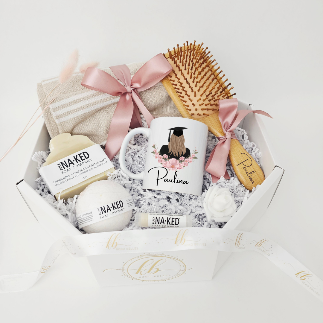 A premium graduation spa care package for her, showcasing a personalized mug with a custom illustration of a graduate from behind surrounded by pink flowers, a name-engraved bamboo brush, a bath bomb, and luxury handmade soap.