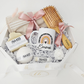 A curated mindful wellness gift box from Koko Belles, showcasing a personalized white mug with a contemporary terracotta and charcoal grey rainbow graphic and the name "Emma". The clean, minimalist package includes a custom-engraved wooden brush, a soft towel with a pink bow, a white rose accent, and natural, soothing bath products by Buck Naked Soap Company.