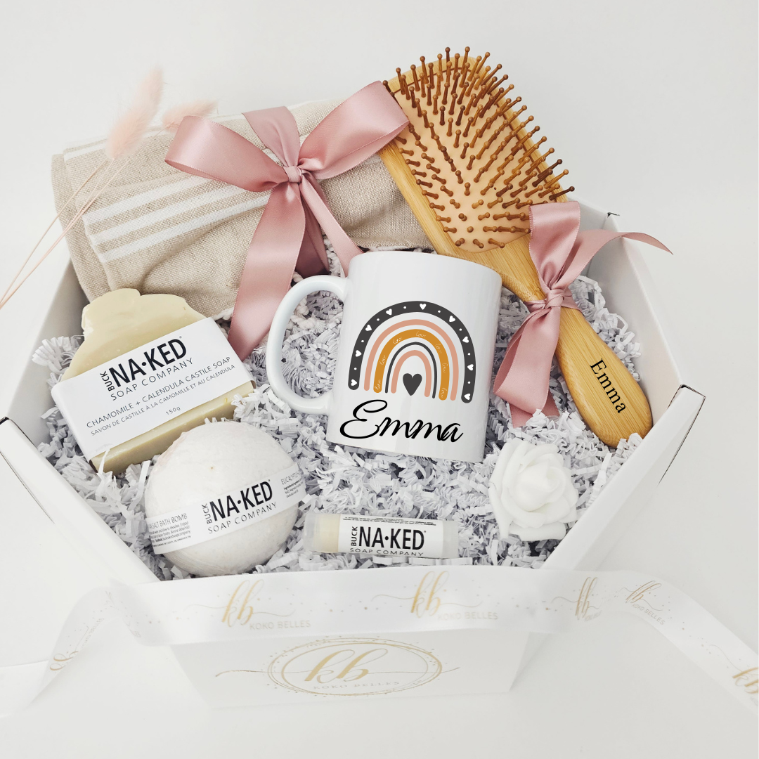 A curated mindful wellness gift box from Koko Belles, showcasing a personalized white mug with a contemporary terracotta and charcoal grey rainbow graphic and the name "Emma". The clean, minimalist package includes a custom-engraved wooden brush, a soft towel with a pink bow, a white rose accent, and natural, soothing bath products by Buck Naked Soap Company.