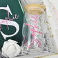 A close-up shot of a Koko Belles gift box featuring a clear glass can tumbler with a bamboo lid and the name "Sophie" personalized in bright pink script. Inside the white and gold-trimmed box, a matching emerald green journal with a large white monogram letter "S" sits next to a delicate white foam rose on a bed of white crinkle paper.