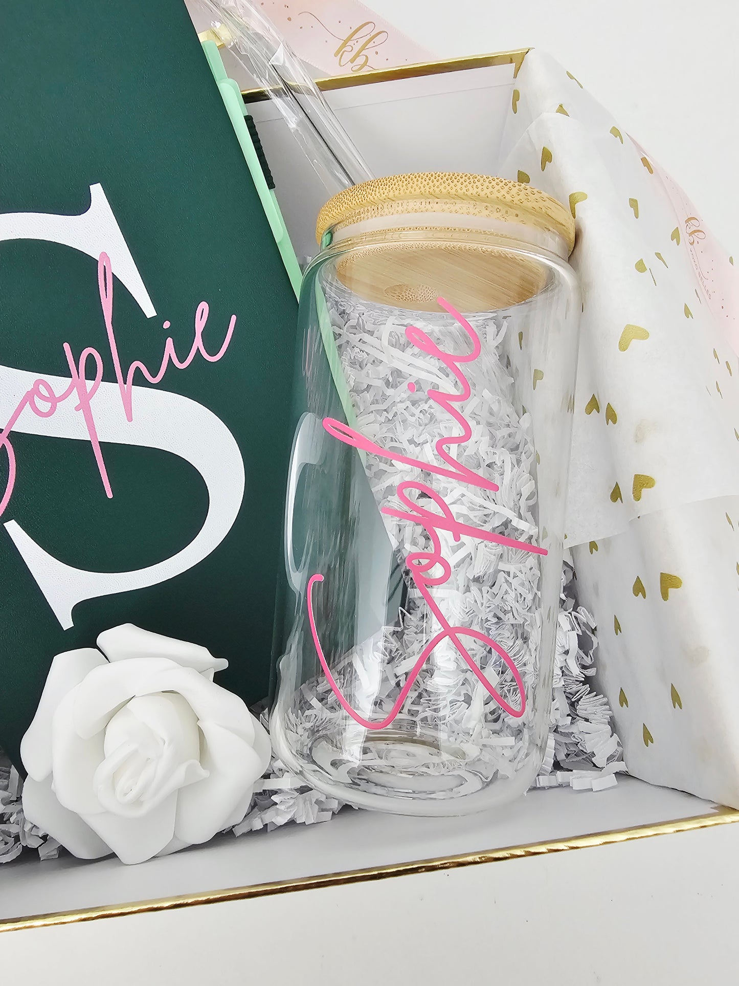 A close-up shot of a Koko Belles gift box featuring a clear glass can tumbler with a bamboo lid and the name "Sophie" personalized in bright pink script. Inside the white and gold-trimmed box, a matching emerald green journal with a large white monogram letter "S" sits next to a delicate white foam rose on a bed of white crinkle paper.