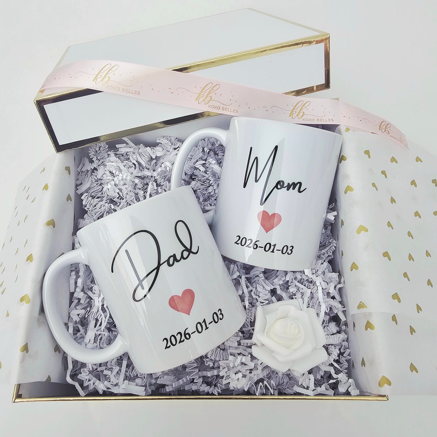 Two white mugs with 'Mom' and 'Dad' personalization, pink hearts, and the date, inside a Koko Belles gift box.