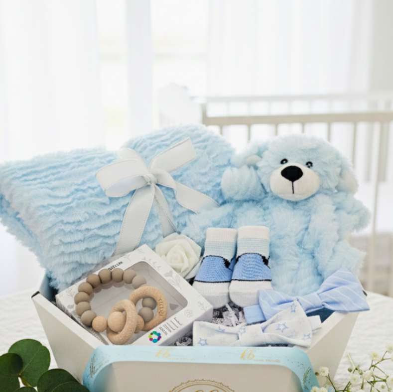 Baby gift set with blue teddy bear, blanket, and socks in a box on a white surface.