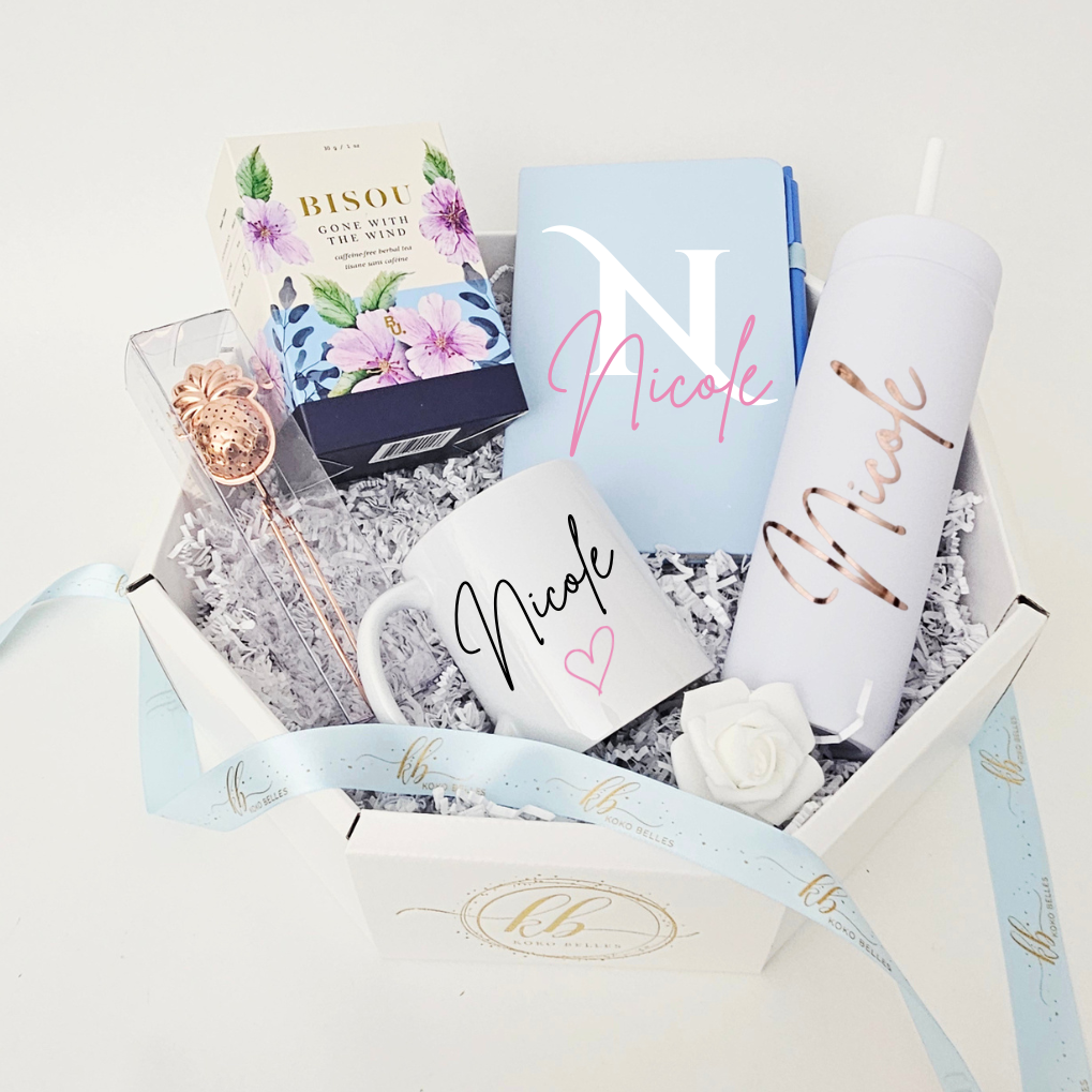 Close-up of a custom light blue notebook and matching pen, paired with a personalized rose gold vinyl skinny tumbler and custom ceramic mug inside a luxury curated gift box by Koko Belles.