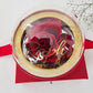 A luxury red gift box with a clear dome bubble lid and gold trim, featuring gold script lettering reading "Ralph and...", filled with deep red roses, accented with a red ribbon and baby's breath flowers on a white background.