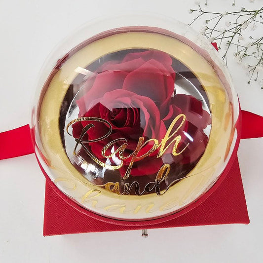 A luxury red gift box with a clear dome bubble lid and gold trim, featuring gold script lettering reading "Ralph and...", filled with deep red roses, accented with a red ribbon and baby's breath flowers on a white background.