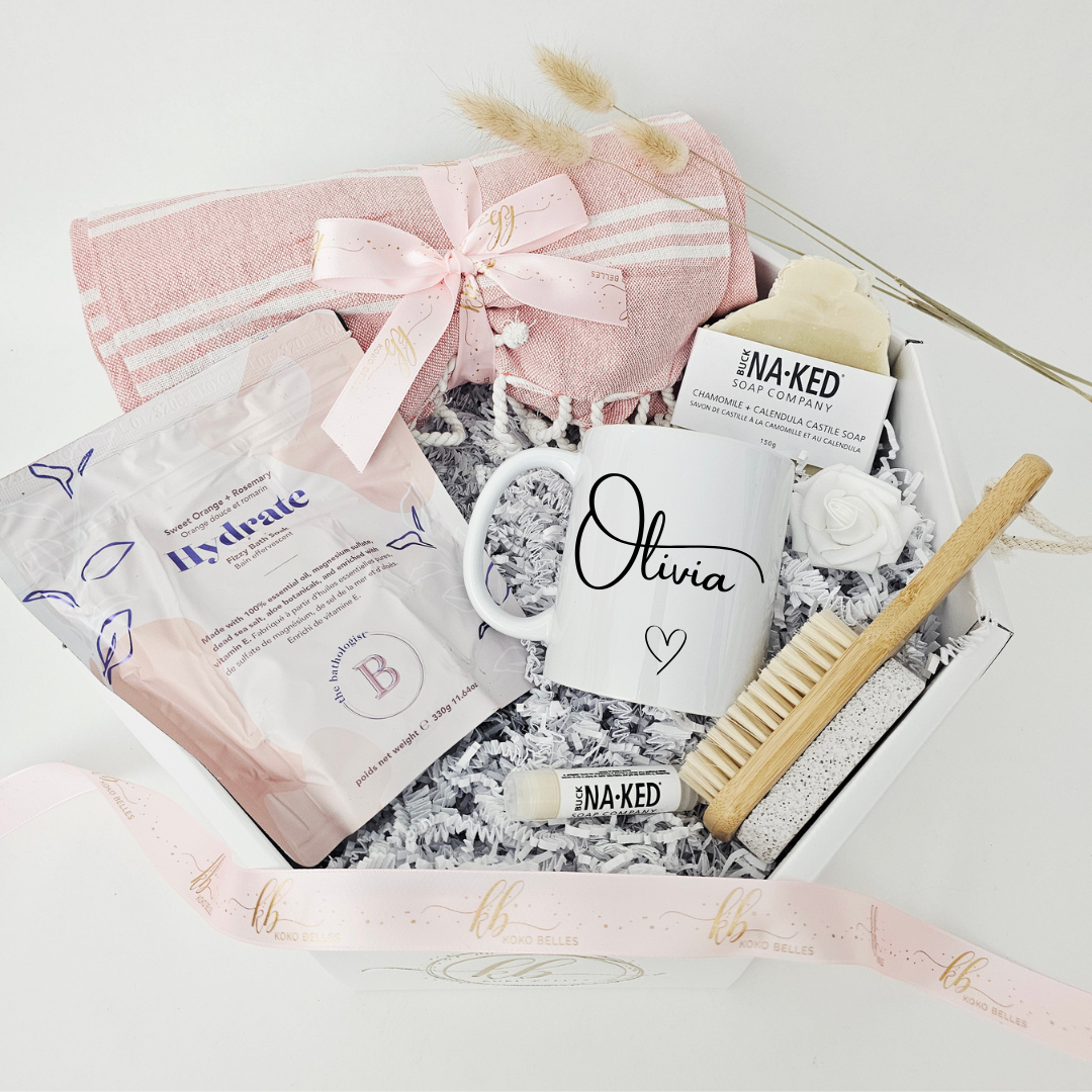 A high-end personalized self-care gift box for women from Koko Belles, elegantly styled in a white octagonal layout. The curated gift package features an artisan soap, a sweet orange and rosemary bath soak, a pink tasseled beach towel, a natural bristle skin brush, and a custom white mug personalized for Olivia.