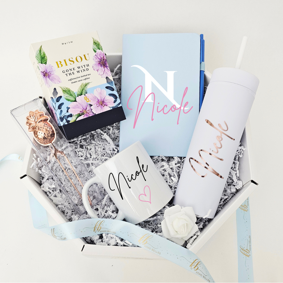 A luxury white octagonal gift box curated with a light blue custom Nicole notebook, a personalized matte white skinny tumbler in rose gold vinyl, a white ceramic coffee mug with a black name and pink heart, Bisou loose leaf tea, and a rose gold pineapple tea infuser.