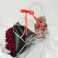 A bouquet of vibrant red roses wrapped in matte black and gold paper, presented inside a clear acrylic tote bag alongside a small plush teddy bear holding a heart.