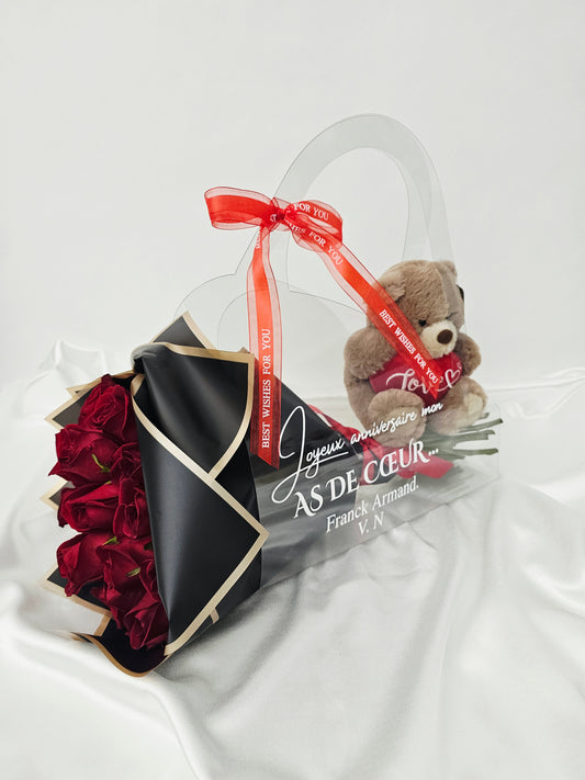 A bouquet of vibrant red roses wrapped in matte black and gold paper, presented inside a clear acrylic tote bag alongside a small plush teddy bear holding a heart.