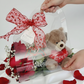 A clear luxury gift tote held by hands, containing red roses and a plush brown teddy bear holding a "Love" heart.