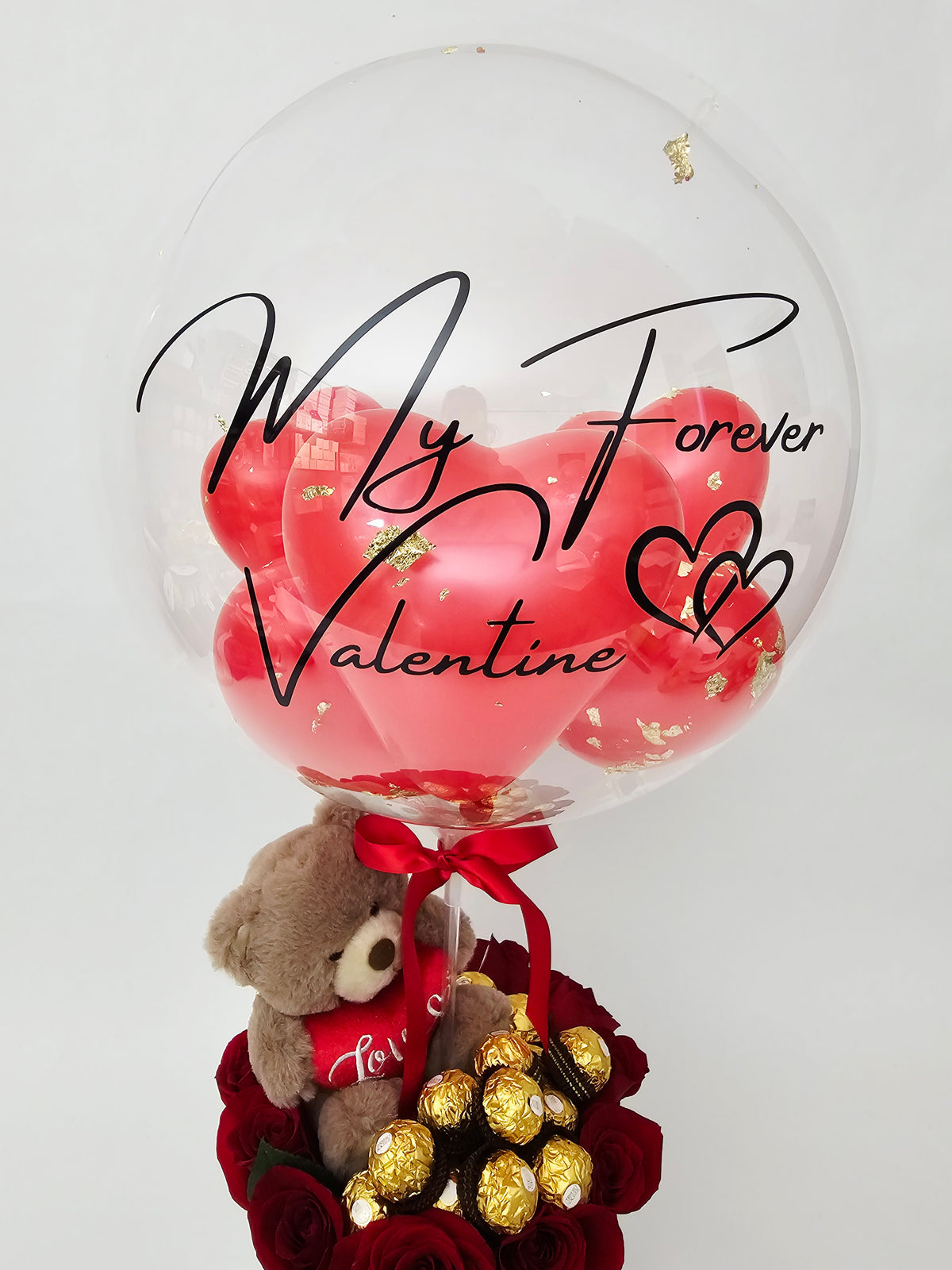 Boîte-cadeau de Saint-Valentin comprenant des roses rouges, des chocolats Ferrero Rocher et un ourson en peluche, surmontée d'un ballon bulle transparent personnalisé « My Forever Valentine ».